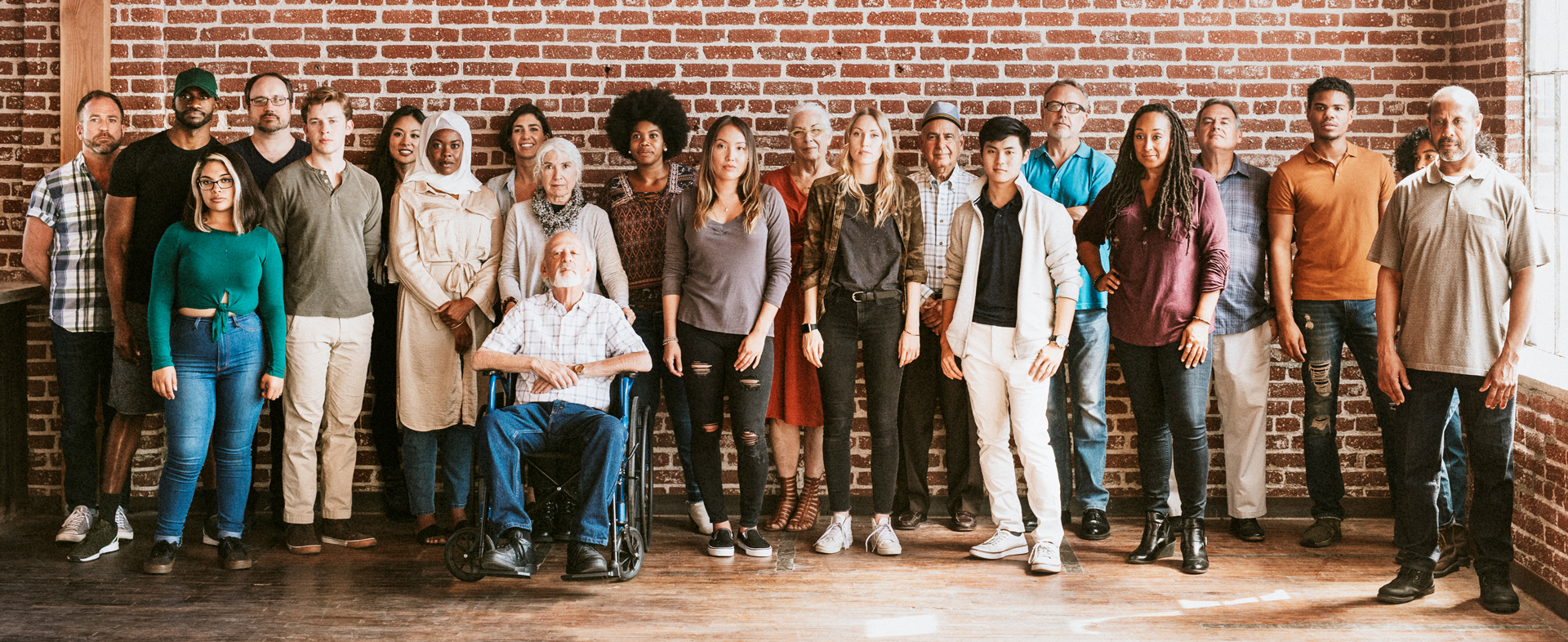 A photo of 21 people positioned toward the camera in front of a brick wall. Most are standing, one is sitting in a wheelchair. They are a wide range of ages, genders, cultures, nationalities.