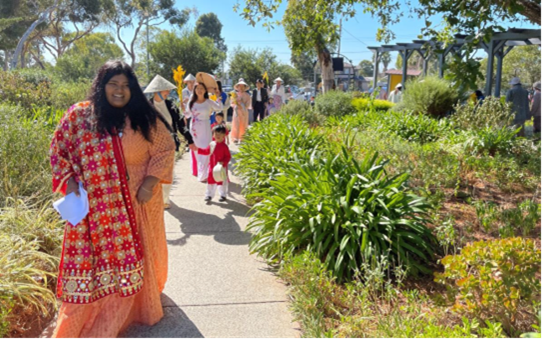 A young woman dressed in a sari walking on a garden path, with a group of adults and children dressed in traditional Vietnamese attire in the background
